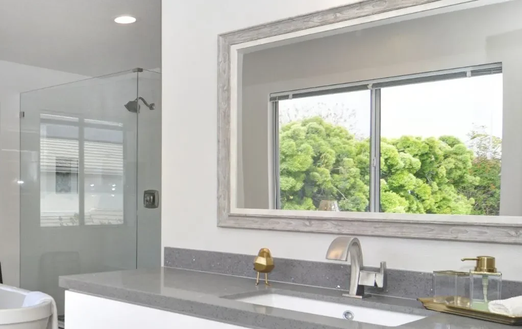 A modern bathroom features a sleek glass shower and a wide mirror, reflecting the lush greenery outside, exemplifying Bayside Builders Group's expertise in home renovations for Bay Area residences.